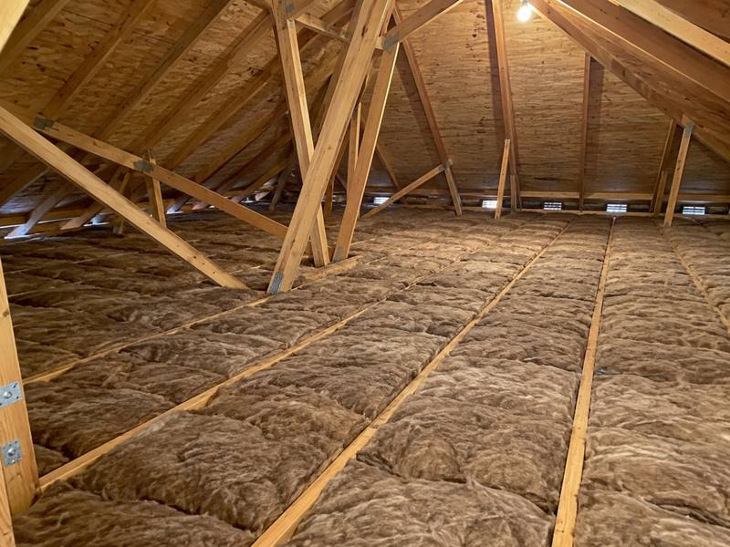 Brown fibreglass batts covering a wide attic floor beneath a web of wooden trusses lit by one bulb