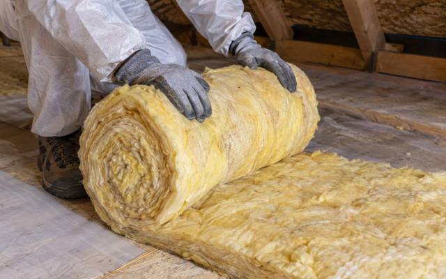 Worker in a protective suit and gloves rolling up a length of yellow fibreglass batt in an attic