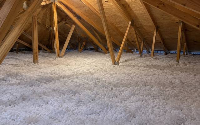 Even layer of white blown-in insulation covering the joists of a long attic lit by a single bulb